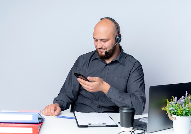 Zadowolony młody łysy mężczyzna call center sobie zestaw słuchawkowy siedzi przy biurku z narzędziami pracy, trzymając i patrząc na telefon komórkowy na białym tle