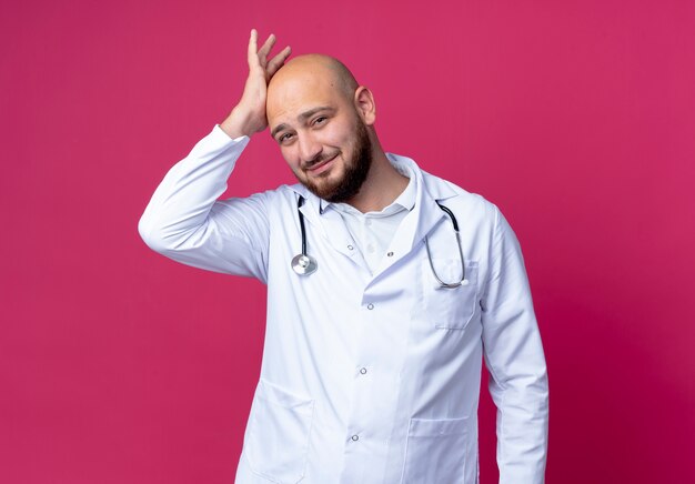 Zadowolony młody mężczyzna łysy lekarz ubrany w szlafrok i stetoskop, kładąc rękę na głowie na białym tle na różowej ścianie