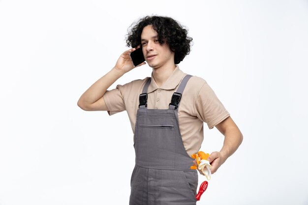 Zadowolony młody mężczyzna pracownik budowlany ubrany w mundur, patrząc na bok rozmawia przez telefon, trzymając w ręku rękawice ochronne z instrumentami budowlanymi w kieszeni na białym tle