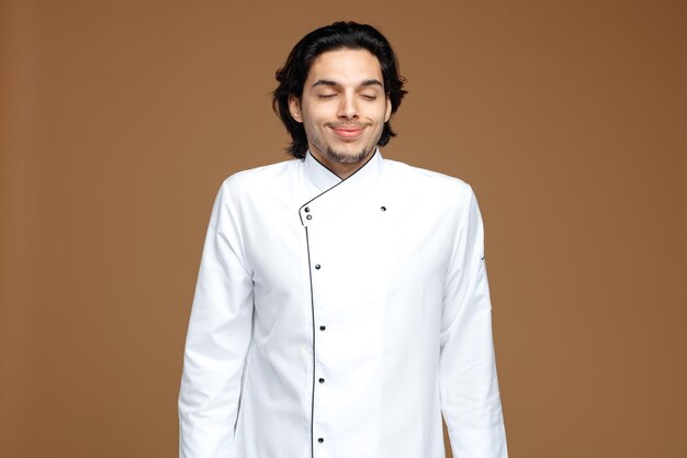 zadowolony młody mężczyzna szef kuchni ubrany w mundur, uśmiechający się z zamkniętymi oczami na brązowym tle