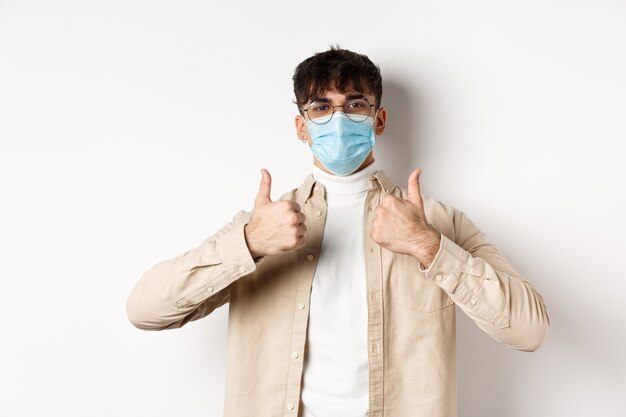 Zadowolony młody mężczyzna w masce na twarz pokazujący kciuki w górę, stosujący środki zapobiegawcze przed koronawirusem...