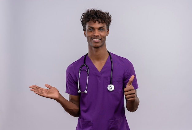 Zadowolony, młody, przystojny, ciemnoskóry lekarz z kręconymi włosami, ubrany w fioletowy mundur ze stetoskopem z kciukami do góry