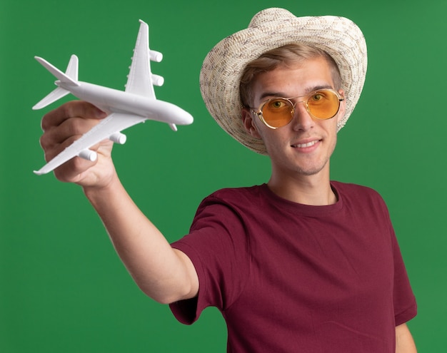 Bezpłatne zdjęcie zadowolony, młody przystojny facet w czerwonej koszuli i okularach z kapeluszem, wyciągając zabawkowy samolot z przodu na białym tle na zielonej ścianie