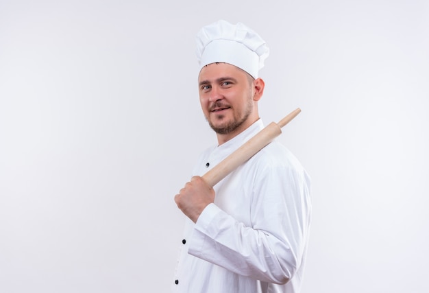 Zadowolony młody przystojny kucharz w mundurze szefa kuchni stojący w widoku profilu i trzymając wałek do ciasta na białym tle na białej przestrzeni