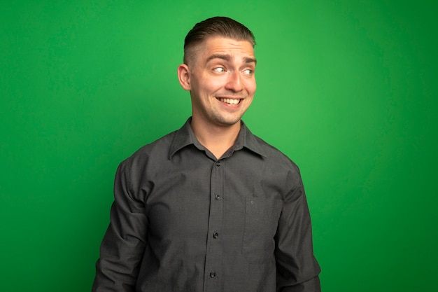 Zadowolony młody przystojny mężczyzna w szarej koszuli patrząc na bok z uśmiechem na twarzy stojącej nad zieloną ścianą