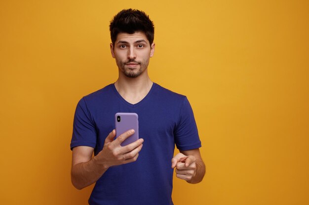 Zadowolony młody przystojny mężczyzna, wskazując i patrząc na kamerę rozciągającą telefon komórkowy w kierunku kamery na żółtym tle