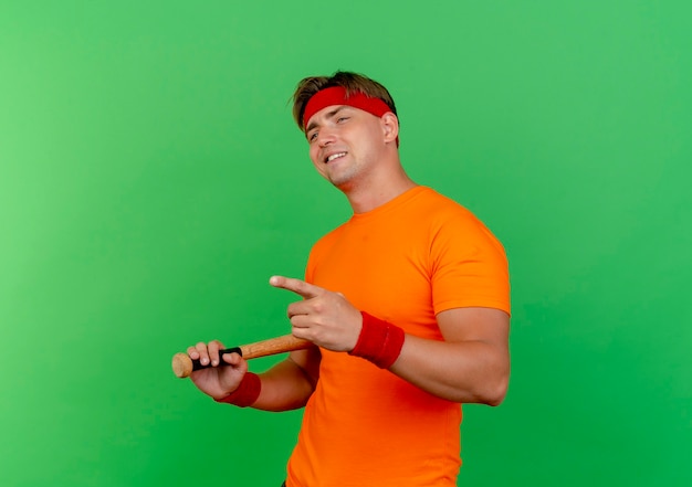 Zadowolony młody przystojny sportowy mężczyzna ubrany w opaskę i opaski na rękę trzyma baseball, patrząc i wskazując prosto nietoperza na zielonej ścianie