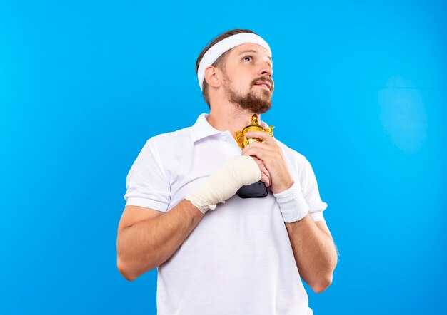 Zadowolony młody przystojny sportowy mężczyzna z opaską na głowę i opaskami na nadgarstkach trzyma puchar zwycięzcy i patrzy w górę z zranionym nadgarstkiem owiniętym bandażem odizolowanym na niebieskiej przestrzeni