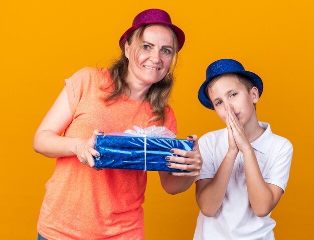 zadowolony młody słowiański chłopiec w niebieskiej imprezowej czapce trzymający się za ręce i stojący z matką w fioletowym imprezowym kapeluszu i trzymającym pudełko na prezenty izolowane na pomarańczowej ścianie z kopią miejsca