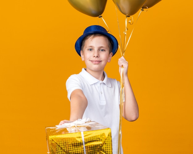 Bezpłatne zdjęcie zadowolony młody słowiański chłopiec z niebieskim kapeluszem strony trzymając balony z helem i pudełko na białym tle na pomarańczowej ścianie z miejsca na kopię