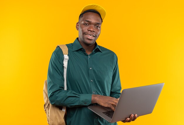 Zadowolony młody student afroamerykański w czapce i plecaku trzymający laptopa