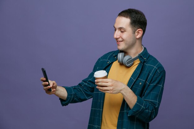 Zadowolony Młody Student Ze Słuchawkami Na Szyi Trzymający Papierową Filiżankę Kawy, Rozciągający Telefon Komórkowy, Biorąc Selfie Na Białym Tle Na Fioletowym Tle