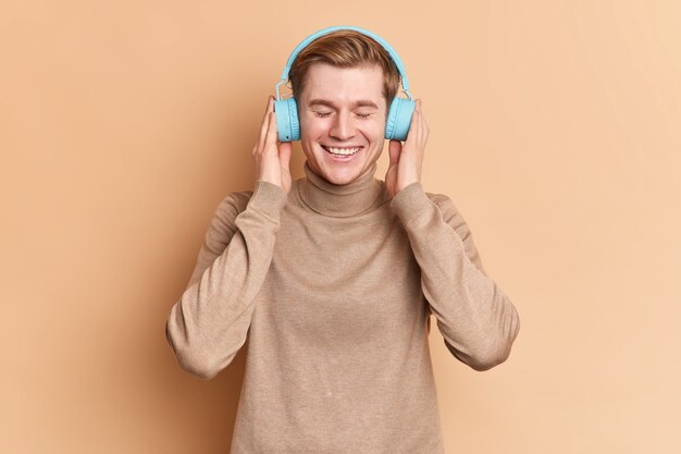 Zadowolony nastolatek relaksuje się z zamkniętymi oczami i słucha ulubionej piosenki przez bezprzewodowe niebieskie słuchawki używa aplikacji muzycznej uśmiecha się z radością nosi swobodne pozy skoczka