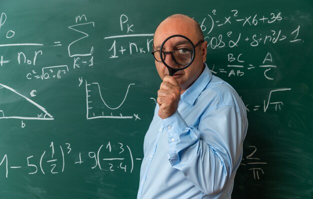 Zadowolony nauczyciel w średnim wieku w okularach stojący przed aparatem patrzącym na tablicę z lupą