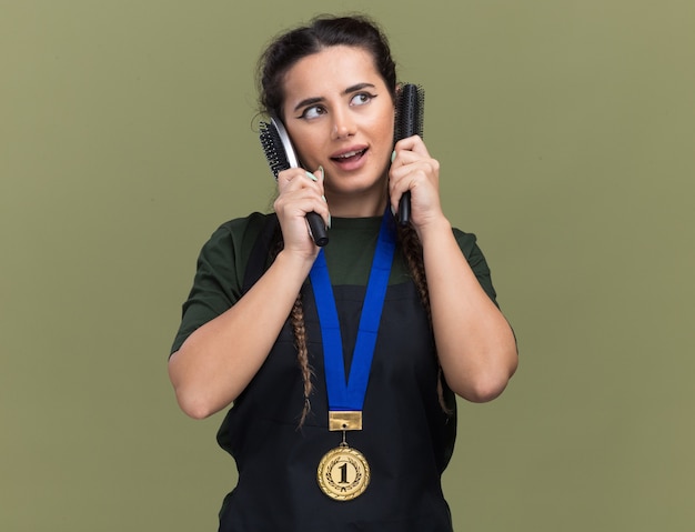 Bezpłatne zdjęcie zadowolony, patrząc na bok młodej fryzjerki w mundurze i medalu, trzymającej grzebienie wokół uszu odizolowanej na oliwkowozielonej ścianie