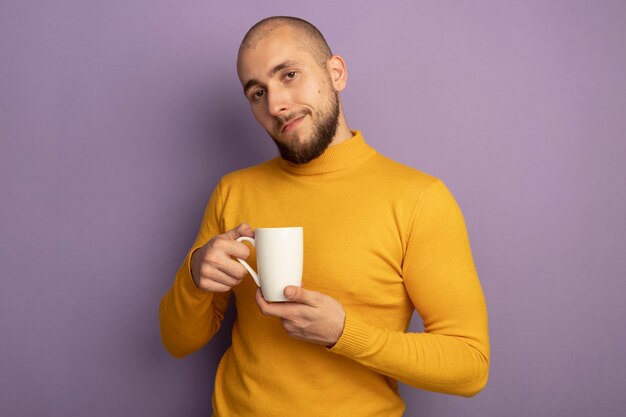 Zadowolony patrząc prosto przed siebie młody przystojny facet trzyma filiżankę herbaty na fioletowym tle