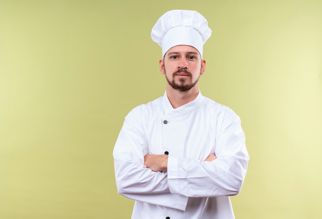 Zadowolony profesjonalny mężczyzna kucharz w białym mundurze i kapelusz stojący z rękami skrzyżowanymi, patrząc pewnie na zielonym tle