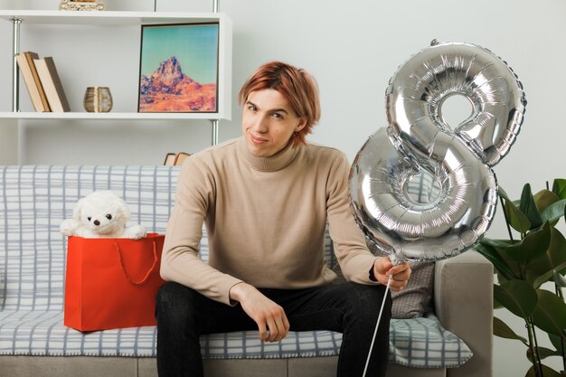 Zadowolony przystojny facet w szczęśliwy dzień kobiet trzymający balon numer osiem siedzący na kanapie w salonie