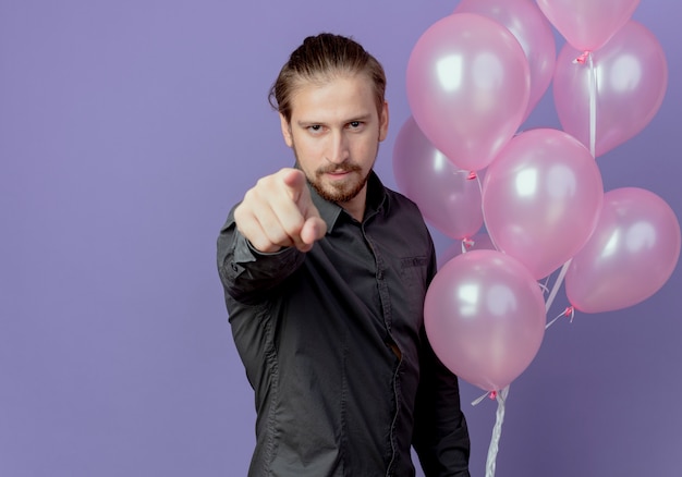 Bezpłatne zdjęcie zadowolony przystojny mężczyzna trzyma balony z helem i punkty na fioletowej ścianie