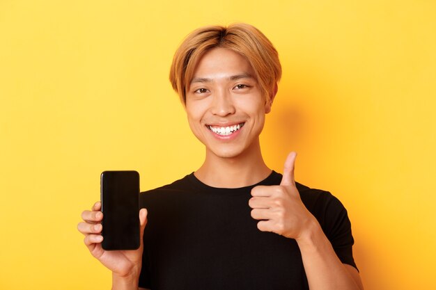 Zadowolony przystojny młody Azjata z jasnymi włosami, pokazujący kciuki do góry na aprobacie i ekranie smartfona, stojący nad żółtą ścianą