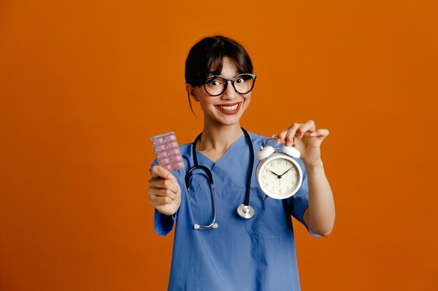 Zadowolony z posiadania budzika z pigułkami młoda lekarka ubrana w jednolity stetoskop fith na białym tle na pomarańczowym tle
