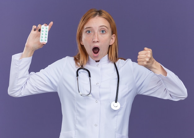Zaimponowana młoda lekarka imbirowa ubrana w szatę medyczną i stetoskop pokazujący paczkę tabletek medycznych do kamery, robiąc silny gest