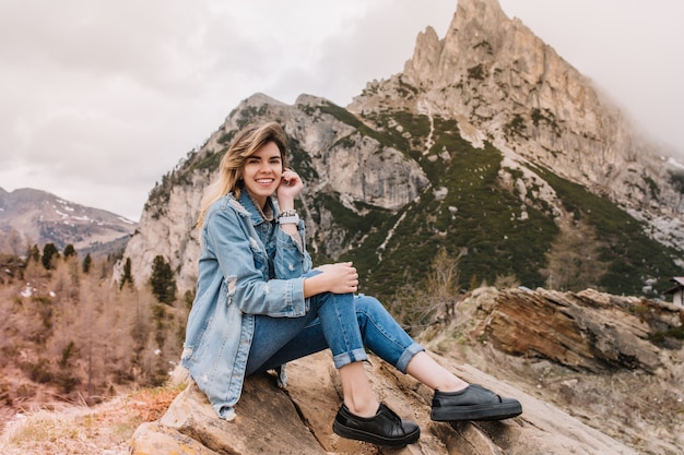 Zainspirowana uśmiechnięta dziewczyna w czarnych skórzanych butach odpoczywa na kamieniu po długim trekkingu i pozuje z przyjemnością