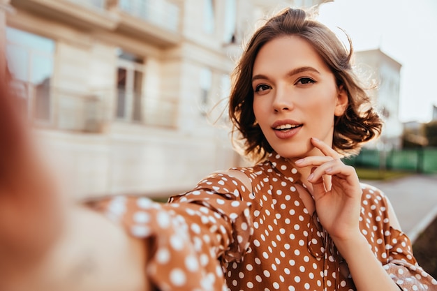 Zainteresowana czarująca kobieta w brązowym stroju robienia selfie. Wspaniała brunetka dziewczyna robi sobie zdjęcie podczas spaceru po mieście.