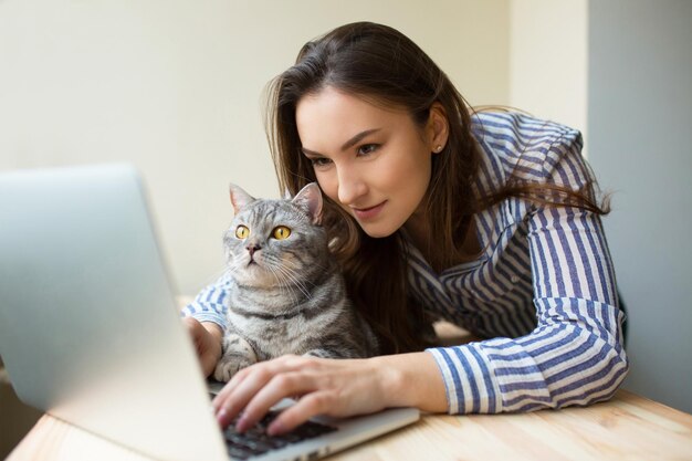 Zainteresowana dziewczyna i jej kot uważnie przyglądają się wyświetlaczowi laptopa. Wesoła dziewczyna trzyma swojego zwierzaka i pracuje przy komputerze jako programista. Dziewczyna i kot korzystający z mediów społecznościowych