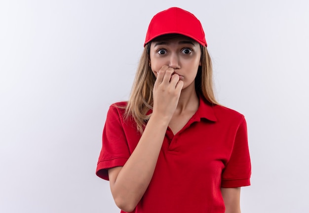 zainteresowana młoda dziewczyna dostawy ubrana w czerwony mundur i czapkę, kładąc rękę na ustach na białej ścianie