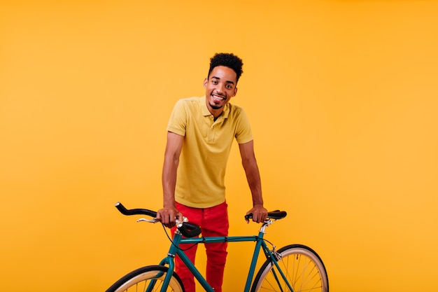 Zainteresowany afrykański model mężczyzna w czerwonych spodniach z uśmiechem. Portret inspirowanego czarnego faceta stojącego w pobliżu zielonego roweru.