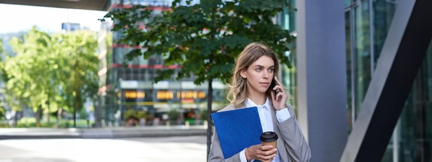 Bezpłatne zdjęcie zajęta bizneswoman pije kawę i rozmawia przez telefon komórkowy, trzymając dokumenty na spacery służbowe w centrum miasta