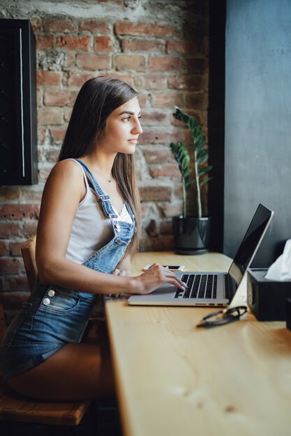 Zajęta młoda modelka siedzi w kawiarni przed oknem, pracuje na swoim laptopie i wypija świeżego drinka