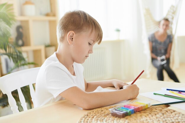 Zajęty chłopiec w wieku kilkunastu lat siedzący w domu z kolorową plasteliną na drewnianym stole, za pomocą ołówka, skoncentrowany na procesie twórczym. Poziomy obraz malarstwa kaukaskiego małego artysty, odrabiania lekcji