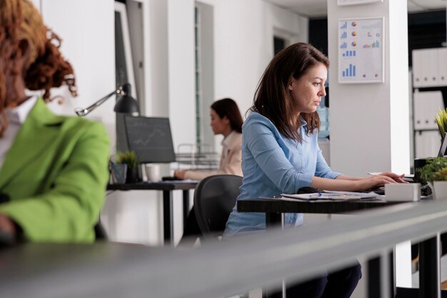 Zajęty pracownik firmy pracujący przy biurku w przestrzeni coworkingowej, kierownik firmy w miejscu pracy, widok z boku. Atrakcyjna poważna kobieta pisząca na laptopie w nowoczesnym biurze, selektywne skupienie