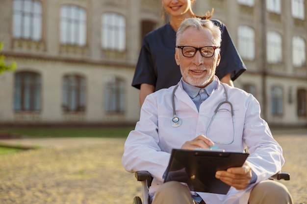 Zajęty starszy niepełnosprawny lekarz płci męskiej na wózku inwalidzkim w fartuchu i okularach z młodą pielęgniarką