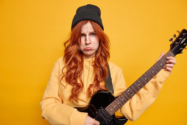Zakłopotana nieszczęśliwa ruda młoda kobieta gra na gitarze elektrycznej basowej, ma smutny wyraz twarzy, nosi czarny kapelusz i casualową żółtą bluzę pozuje w pomieszczeniu. Niezadowolony rocker z instrumentem muzycznym