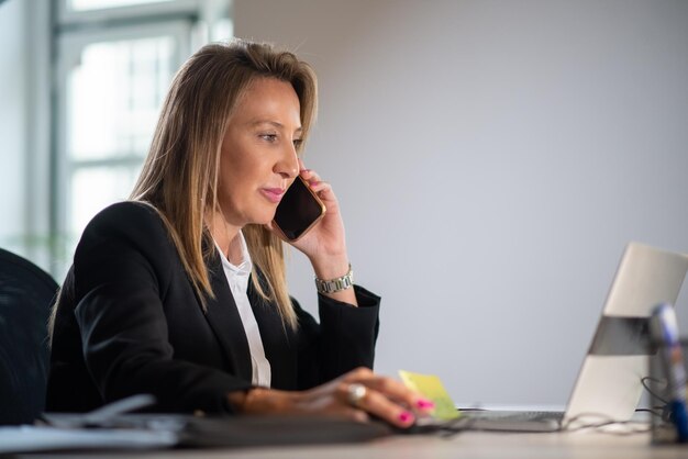 Zakończenie bizneswoman z telefonem. Poważna kobieta siedzi przy stole, patrząc na laptopa za pomocą telefonu, rozmawiając z partnerami o nowych projektach i wsparciu finansowym. Koncepcja rozwoju biznesu i finansów