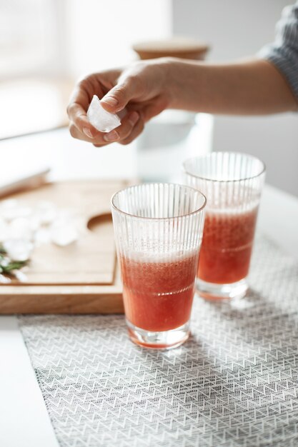 Zakończenie kobiet ręki stawia lodowe kawałki w szkłach z grapefruitowym detox zdrowym smoothie z bliska.