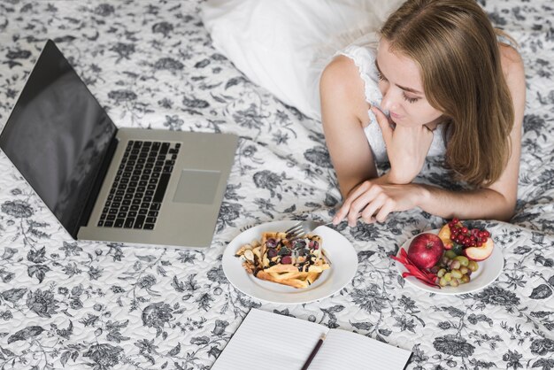 Zakończenie kobieta patrzeje laptopu łasowania gofr z jagodami na kwiecistym łóżku