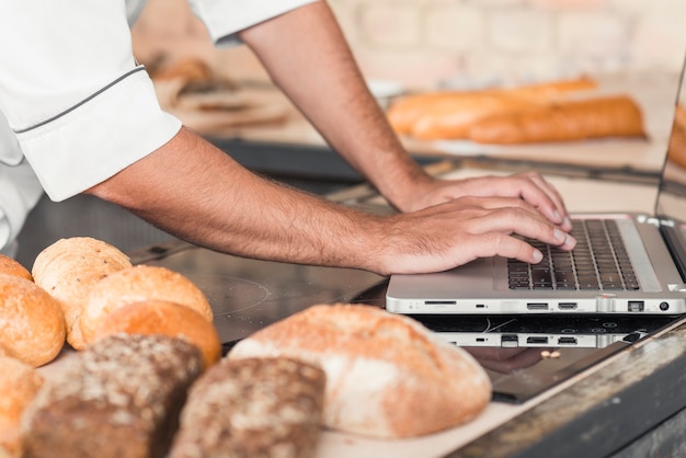 Bezpłatne zdjęcie zakończenie męska piekarz ręka używać laptop na kuchennym worktop z chlebami