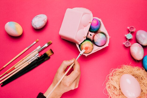 Zakończenie osoby ręka maluje jajko z farby muśnięciem w kartonie na różowym tle