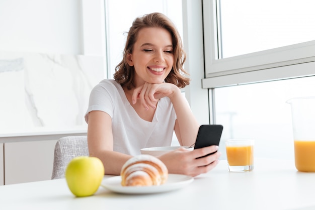 Zakończenie portret szczęśliwa brunetki kobieta używa telefon komórkowego podczas gdy mieć śniadanie przy białą kuchnią