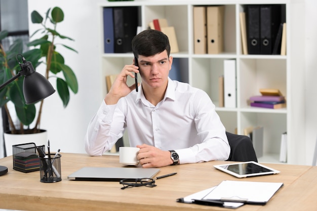 Zakończenie poważny młody biznesmen trzyma filiżankę kawy opowiada na telefonie komórkowym przy miejscem pracy