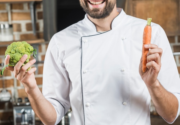 Bezpłatne zdjęcie zakończenie uśmiechnięta szefa kuchni mienia marchewka i brokuły w jego ręce