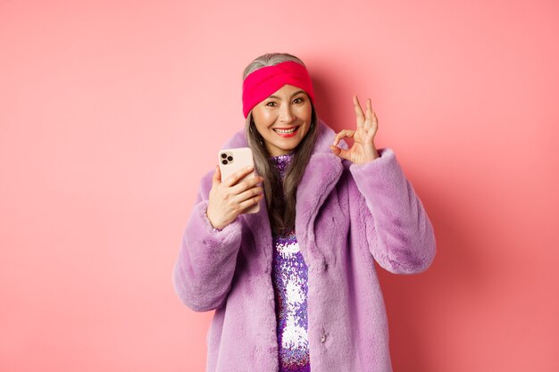 Zakupy online i koncepcja mody. Stylowa azjatycka starsza kobieta pokazująca dobry znak i trzymająca telefon komórkowy, polecająca sklep internetowy, różowe tło