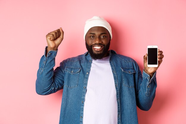 Zakupy online i koncepcja technologii. Wesoły Murzyn radujący się i pokazujący ekran mobilny, podnoszący zadowoloną rękę, triumfujący stojąc na różowym tle