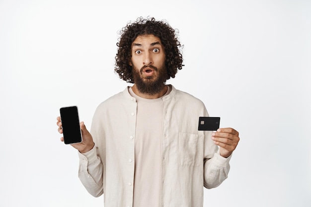 Zakupy online Młody zdumiony arabski facet pokazujący ekran telefonu komórkowego i kartę kredytową, wpatrujący się w kamerę białe tło