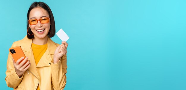 Zakupy online Stylowa młoda Azjatka w okularach przeciwsłonecznych pokazująca kartę kredytową i używająca smartfona płacącego w internecie, dokonującego zakupu stojącego na niebieskim tle