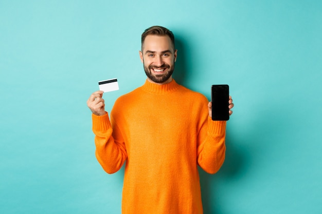 Zakupy online. Szczęśliwy atrakcyjny facet pokazujący ekran telefonu komórkowego i kartę kredytową, uśmiechnięty zadowolony, stojący nad jasną turkusową ścianą